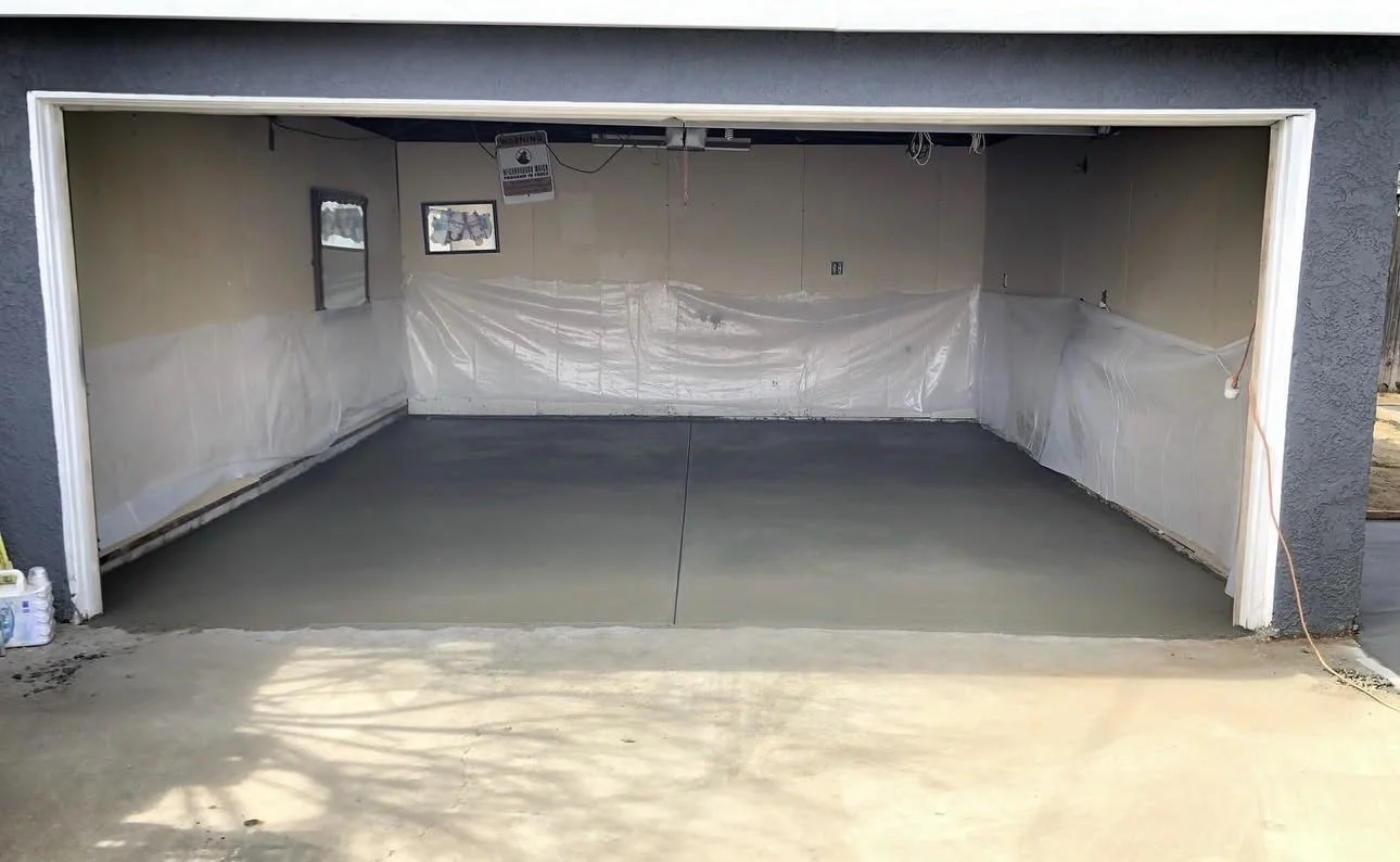 Empty garage with newly poured concrete floor, partially covered with white plastic sheeting, with two small windows and construction materials on the side.
