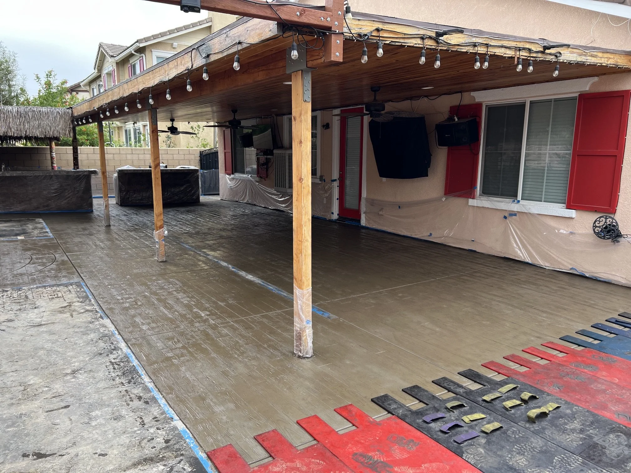 A backyard patio area under construction with a wooden deck and a wood-covered pergola, decorated with string lights. The space features red shutters on windows, ceiling fans, and various construction materials and equipment.