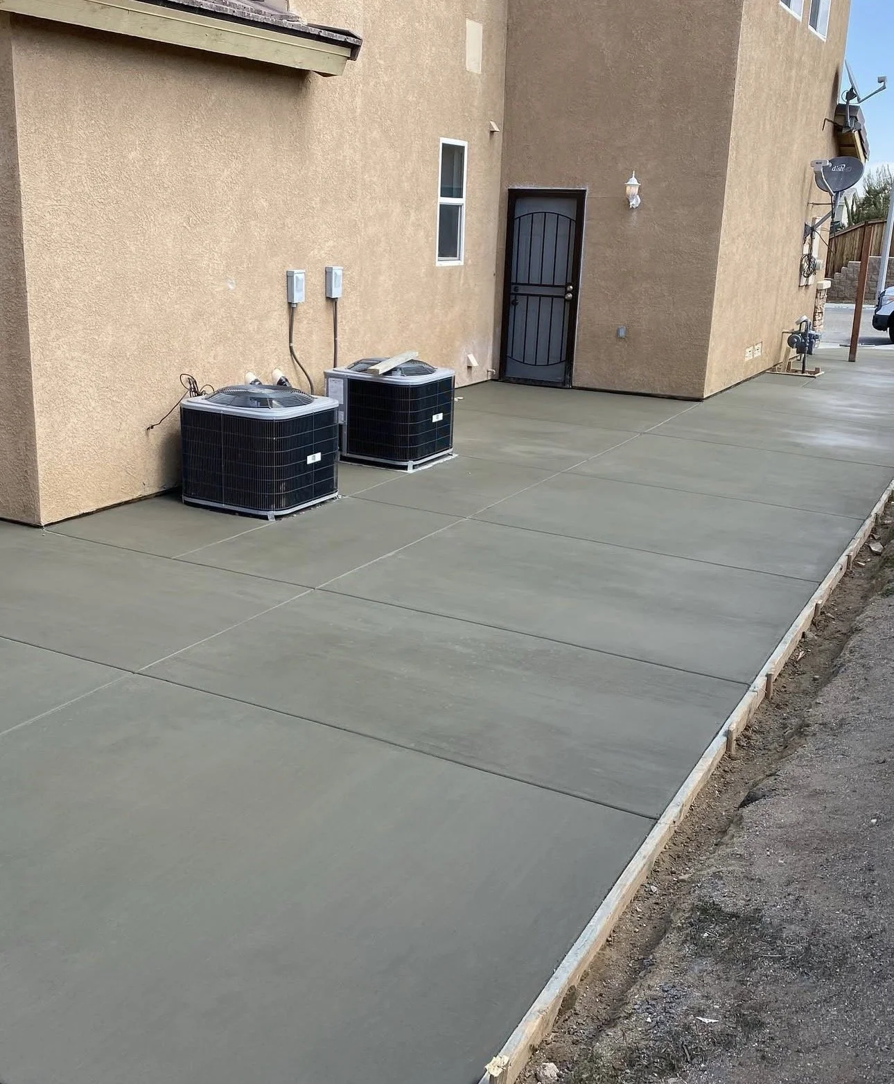 A recently paved concrete patio exterior of a building with two air conditioning units, a door, and exterior lighting, with a narrow dirt border along the edge.