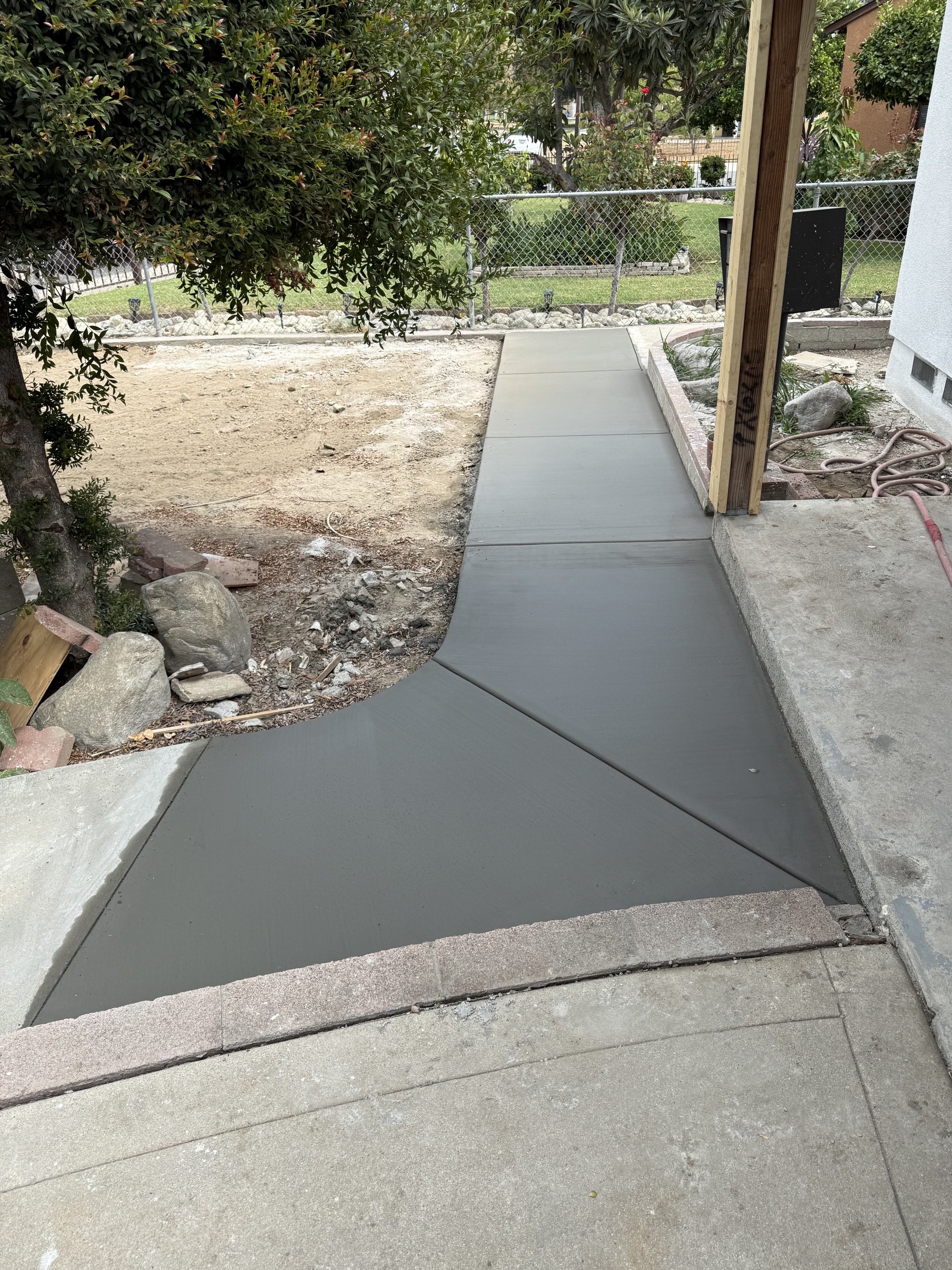 Newly poured concrete sidewalk in front of a house, with landscaping and trees in the background.