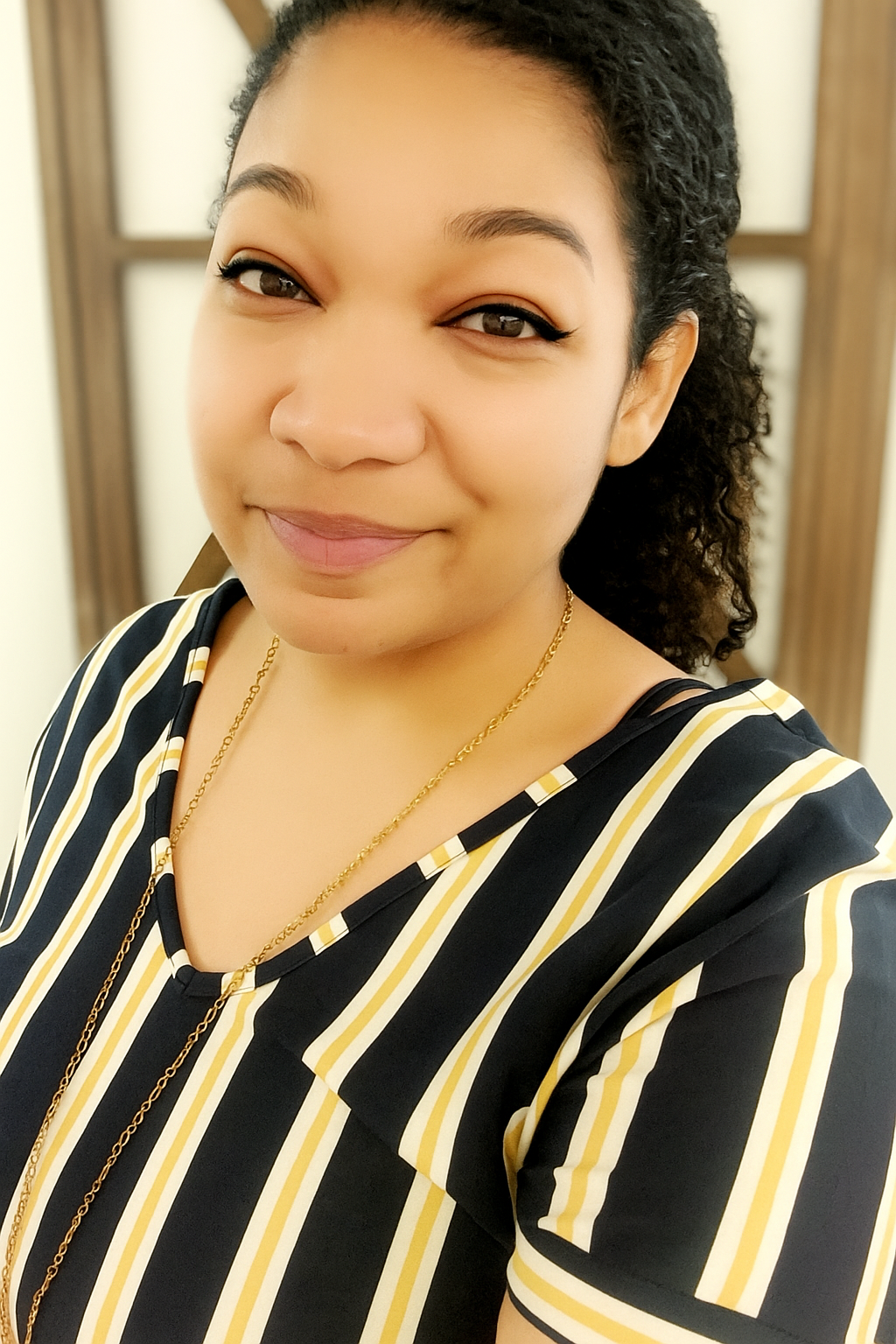 Close-up portrait of a woman with dark curly hair, wearing a black and yellow striped top and gold jewelry, smiling softly in front of a wooden framed mirror.