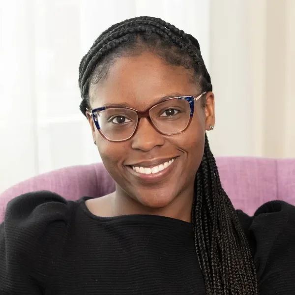A woman with glasses and braided hair smiling while sitting on a purple chair.