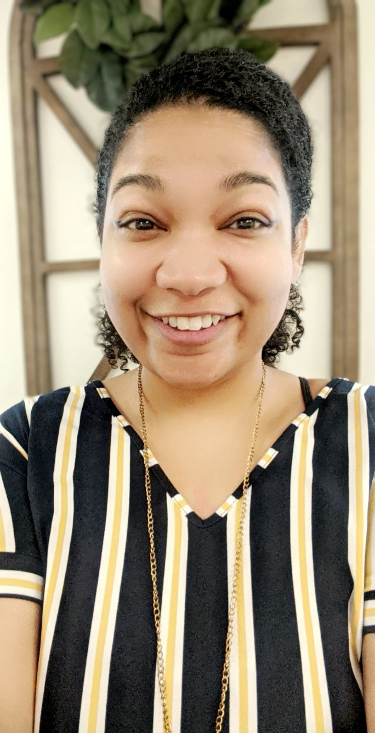 A smiling woman with short curly hair, wearing a black, yellow, and white striped top, a gold necklace, and a gold chain, standing in front of a wooden lattice with green plants in the background.