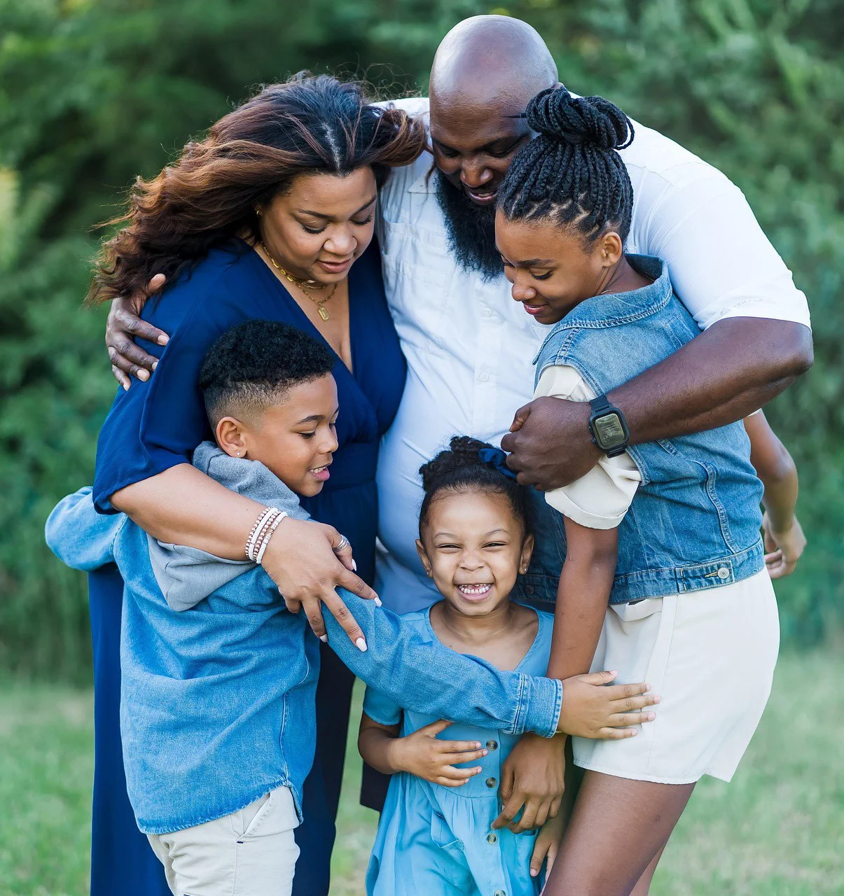 A family of six, including two parents and four children, sharing an embrace outdoors on a grassy area with green trees in the background.