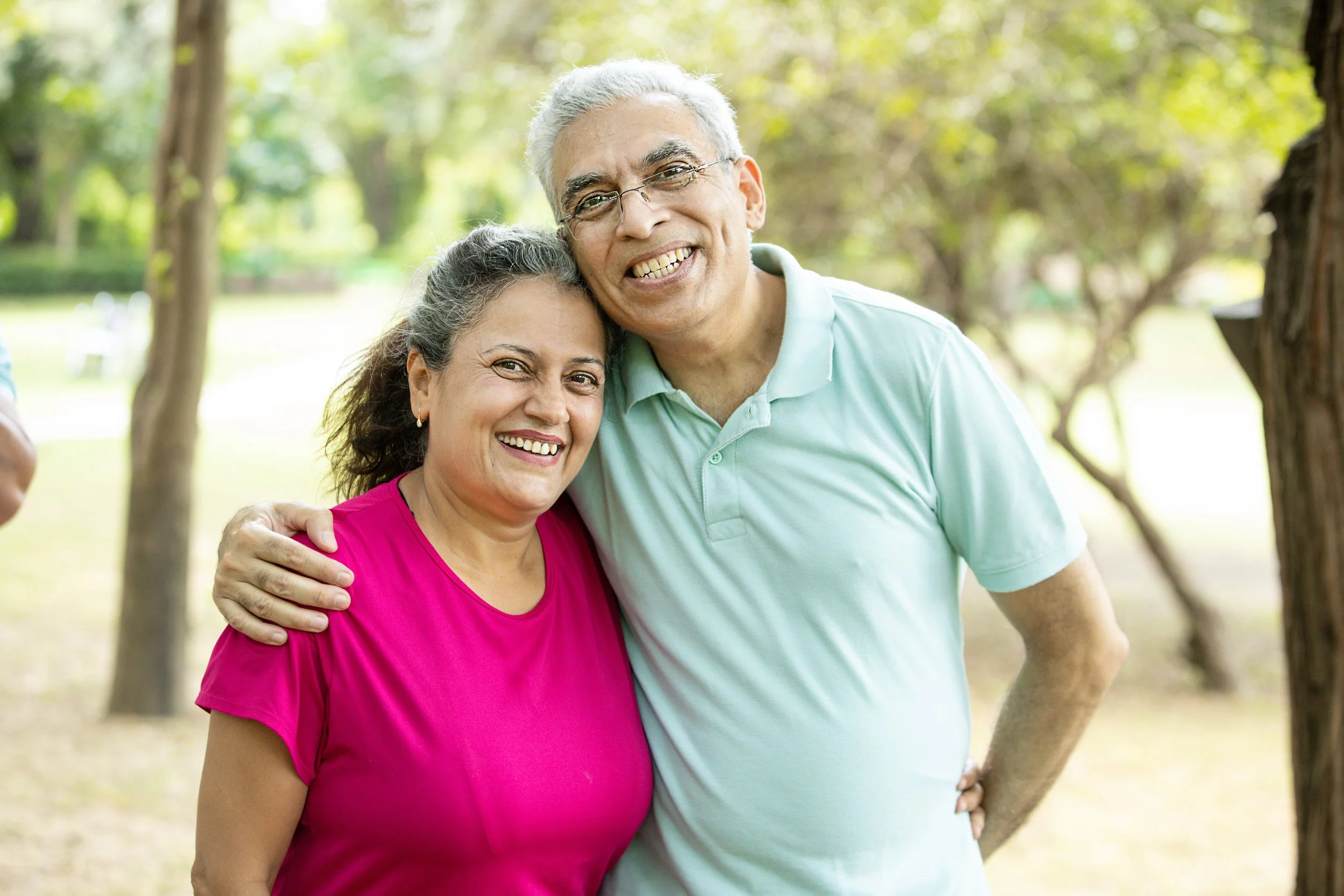 An elderly couple hugging and smiling in a park with green trees in the background.