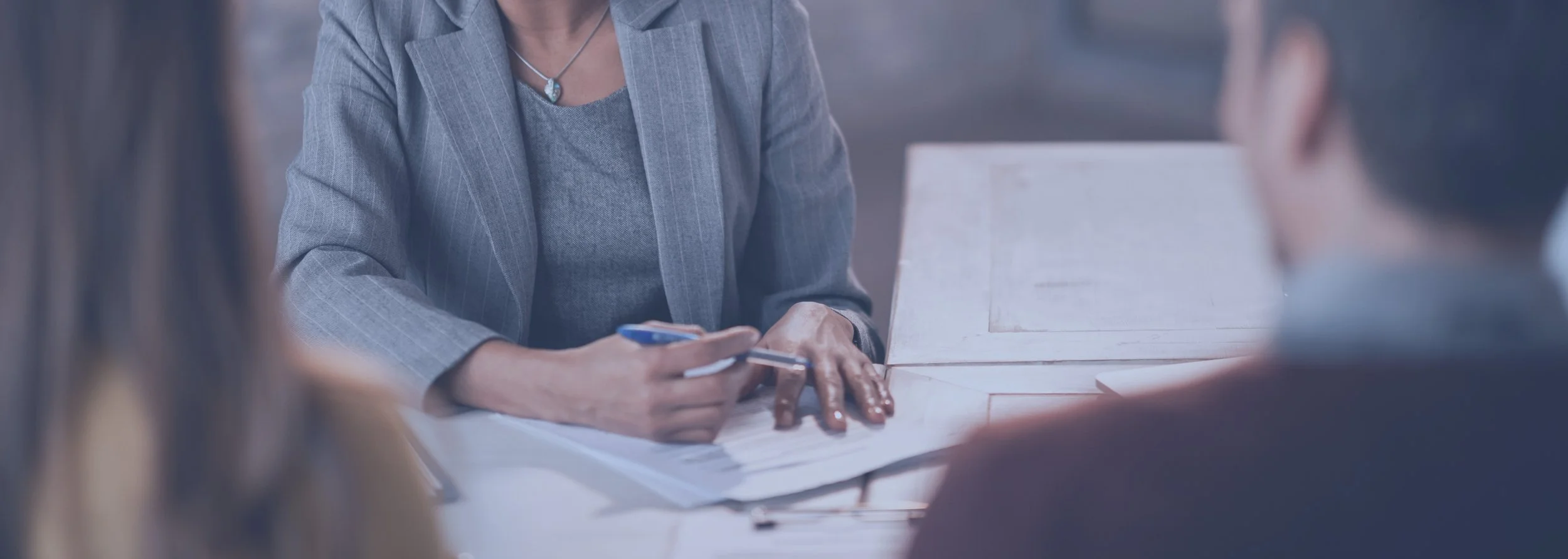 A woman in a gray blazer and matching top sitting at a table during a meeting, holding a pen and looking at documents, with other people around her.
