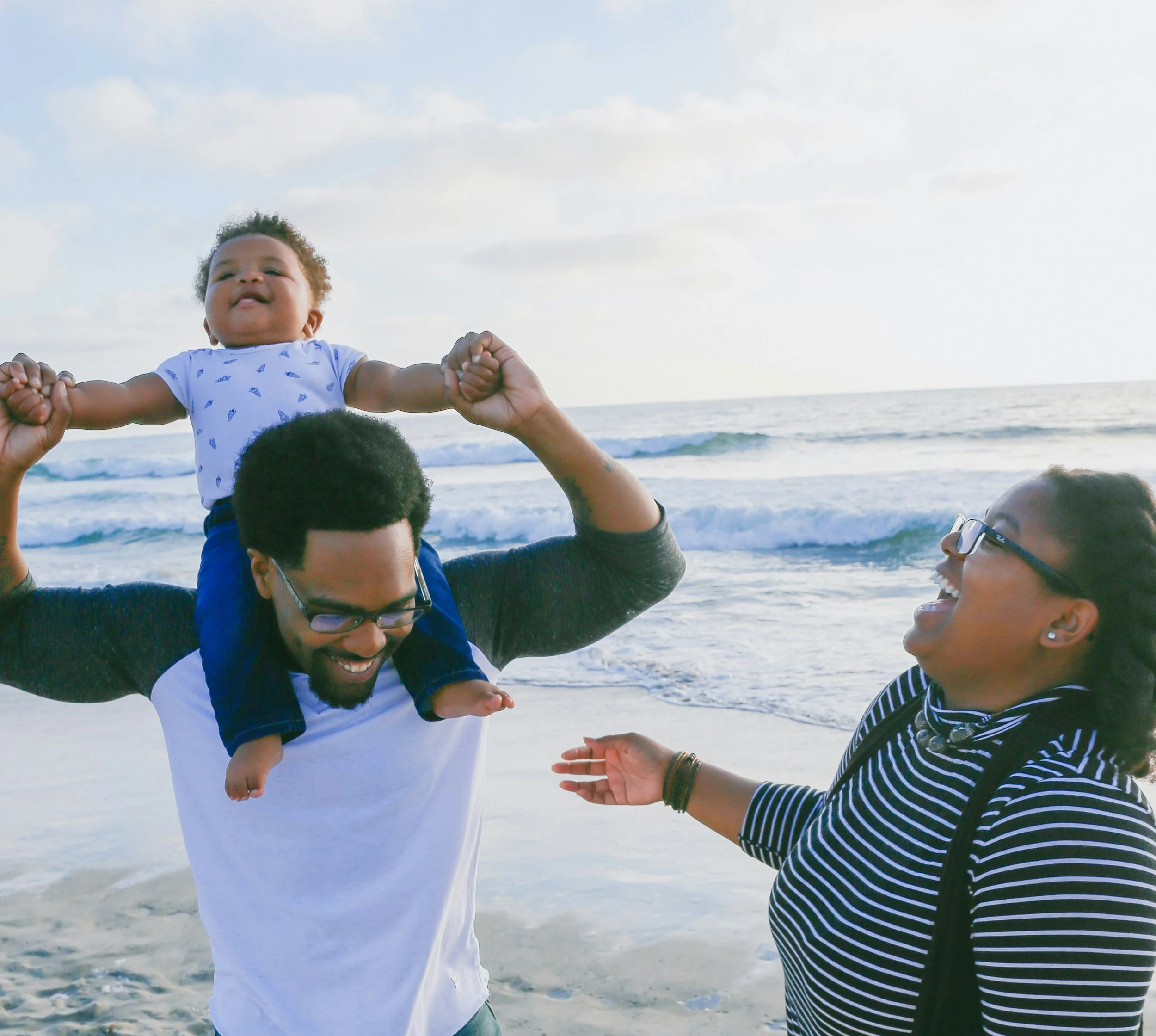 A baby being lifted by adults at the beach with the ocean and sky in the background.