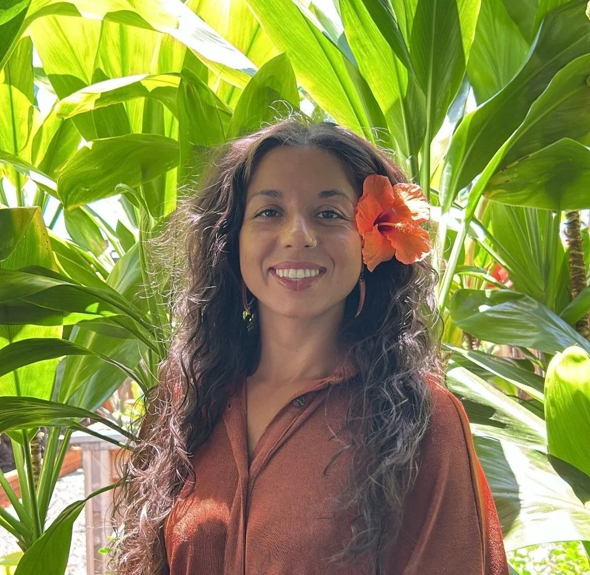A woman with long, curly dark hair and a large orange flower in her hair, smiling, surrounded by lush green tropical plants.