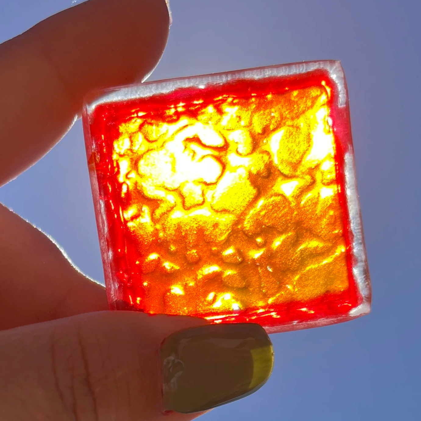 A small translucent orange and red square tile held up to the sky with the sunlight shining through the back