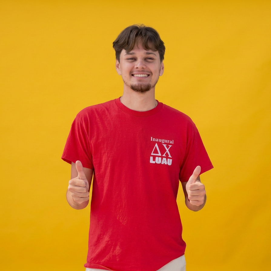 A young man with brown hair and a goatee smiling and giving two thumbs up while standing against a yellow background. He is wearing a red T-shirt with the words "Inaugural AX LUAW" printed on it.
