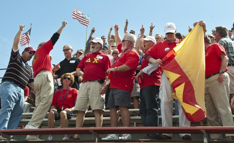 Group of people cheering and waving flags, some wearing red shirts, at an outdoor event under a clear blue sky.