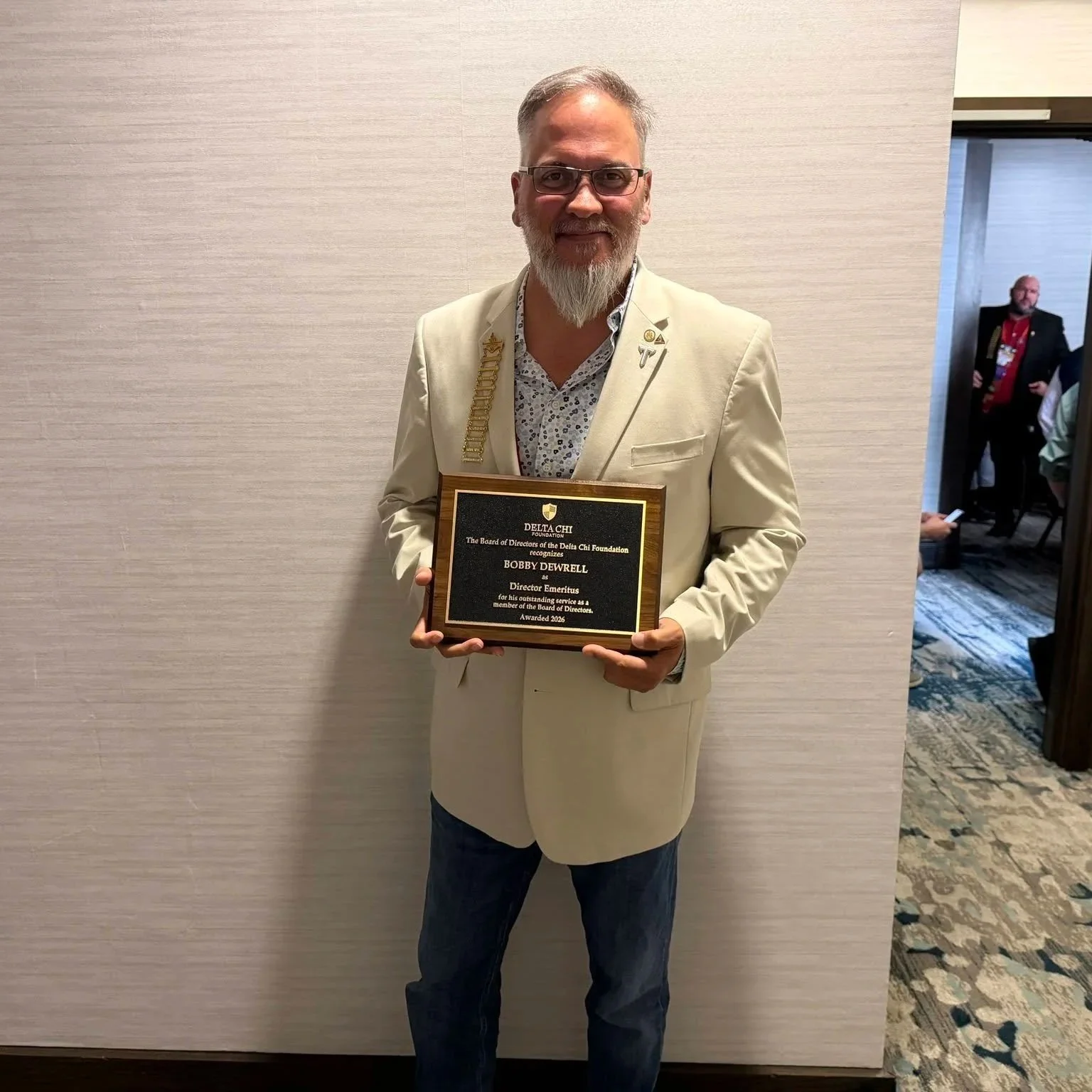 A man with glasses and a beard wearing a cream-colored suit holding a plaque, standing in front of a light-colored wall at an awards event.
