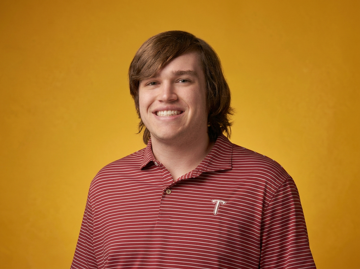 Young man with medium-length brown hair, smiling, wearing a maroon striped collared shirt with a small white 'T' logo on the chest, standing against a plain yellow background.