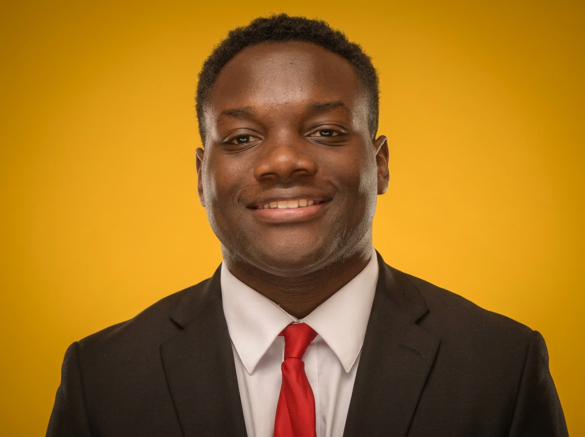 A man in a black suit, white shirt, and red tie smiling against a yellow background.