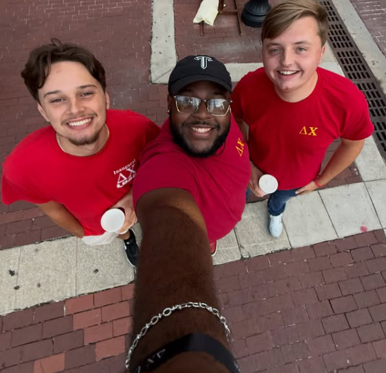 Three young men taking a selfie from above on a brick sidewalk, all wearing red T-shirts, with two holding cups and smiling.