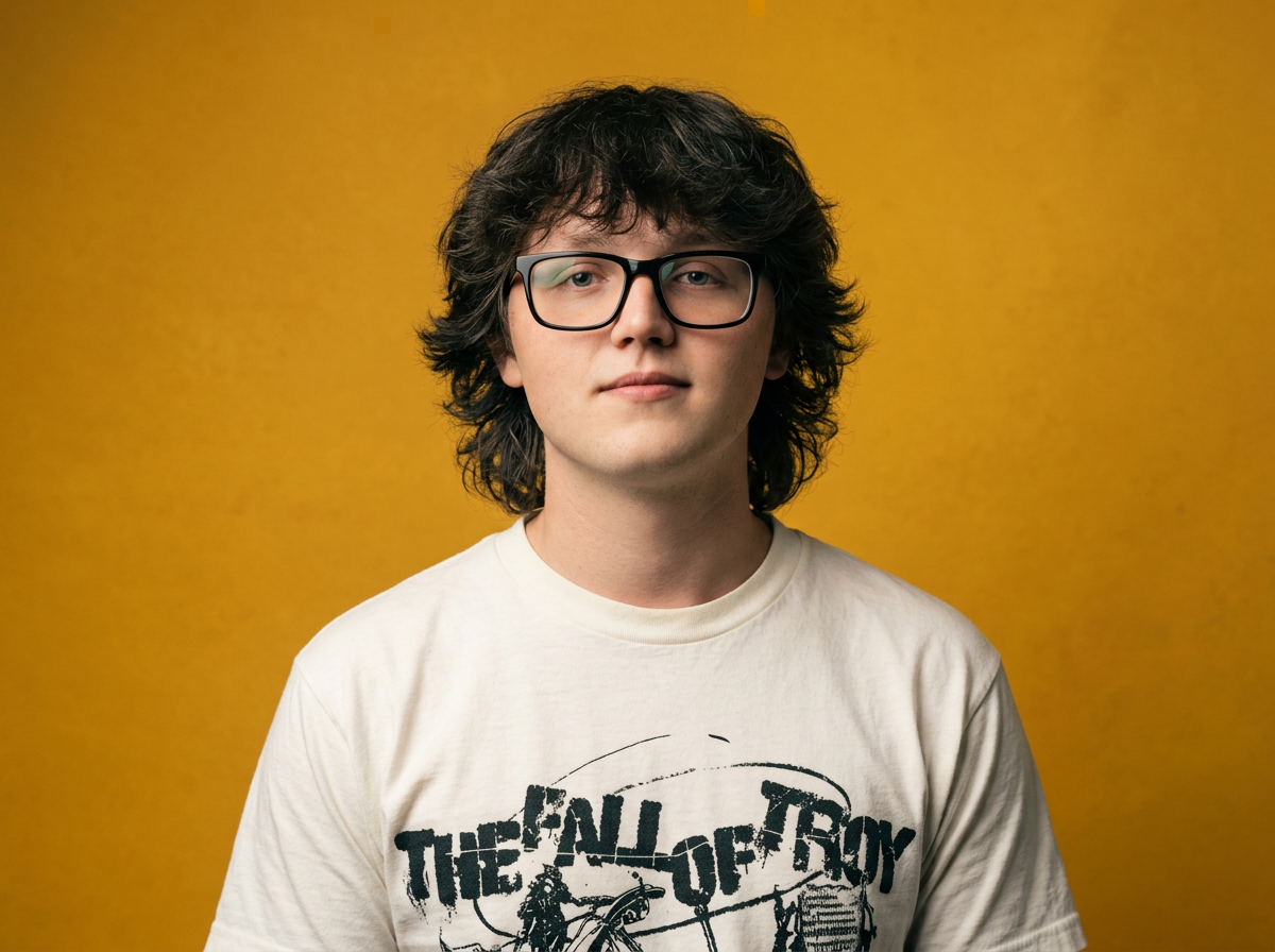 Young man with glasses and wavy hair standing against a yellow background, wearing a white T-shirt with text and graphic design.