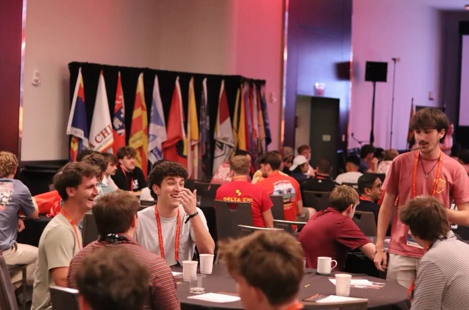 Group of teenagers at an indoor event, seated at round tables with cups, with flags in the background, some wearing red shirts and lanyards, engaged and smiling.