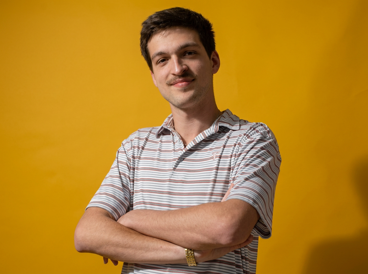 A young man with dark hair wearing a striped polo shirt and a gold watch, standing with arms crossed in front of a yellow background.