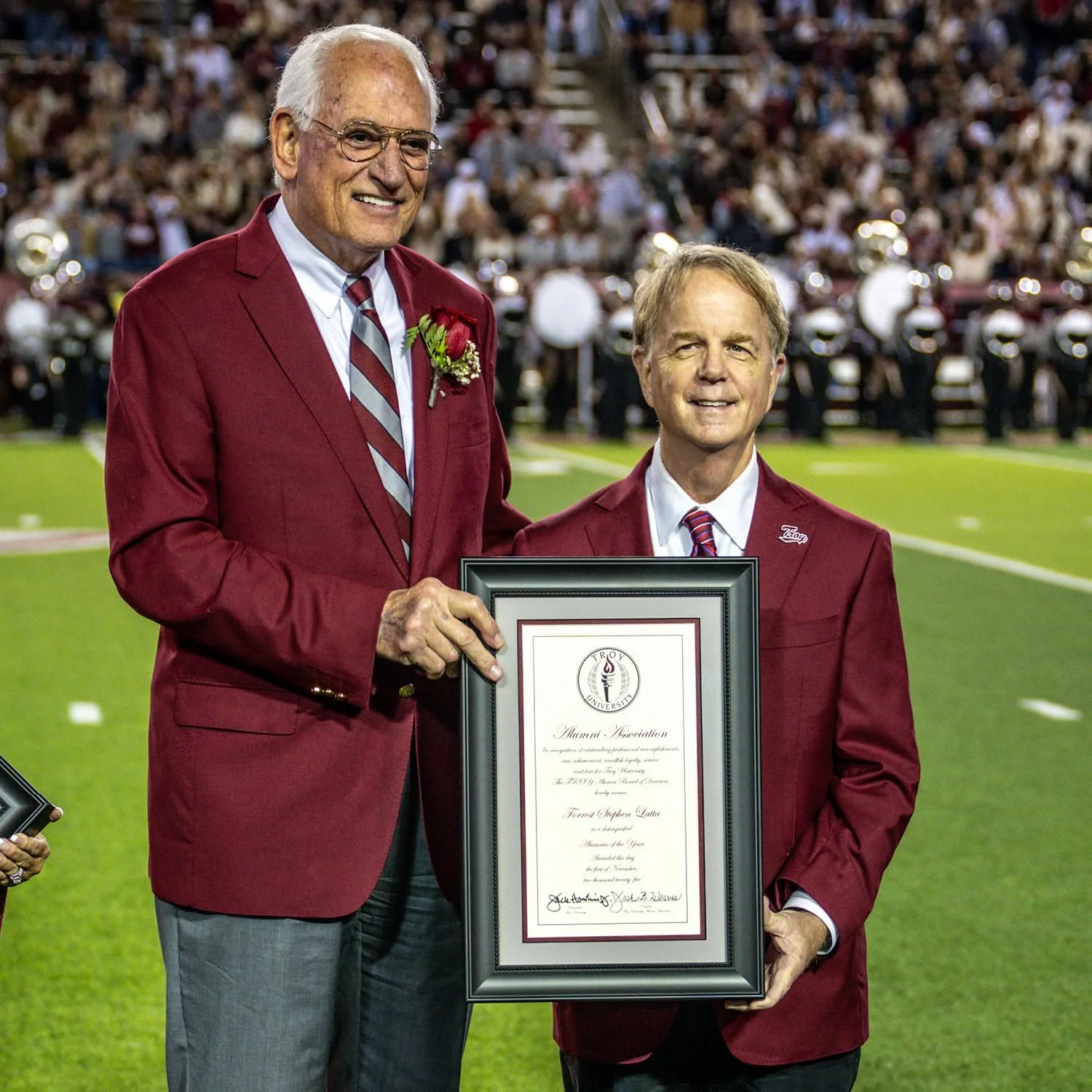 Two men in red blazers standing on a football field, with one holding a framed certificate. The man on the right is smiling and the man on the left is also smiling. In the background, there are spectators and a marching band.