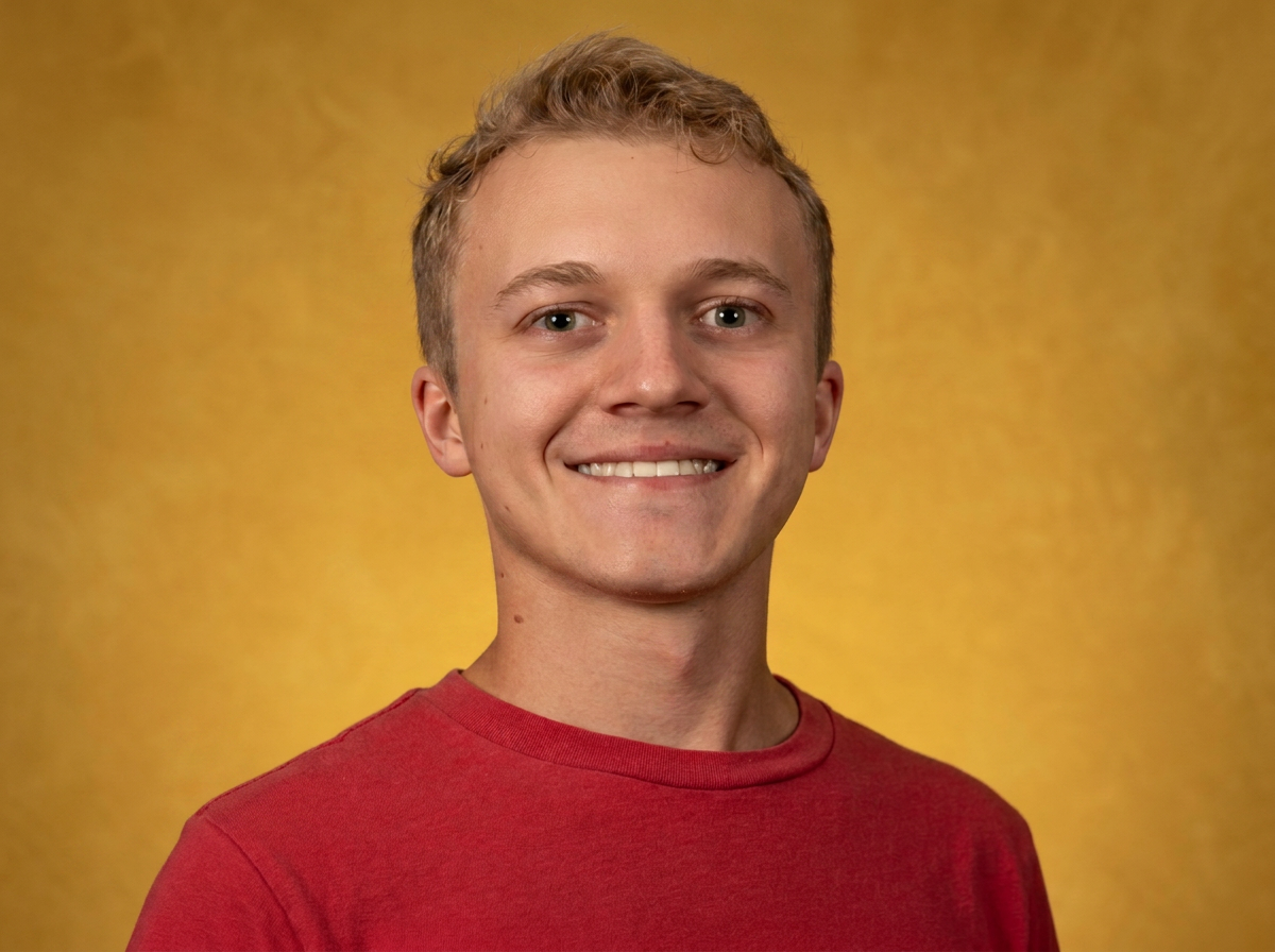 A young man with short, curly blonde hair is smiling and wearing a red shirt against a yellow background.
