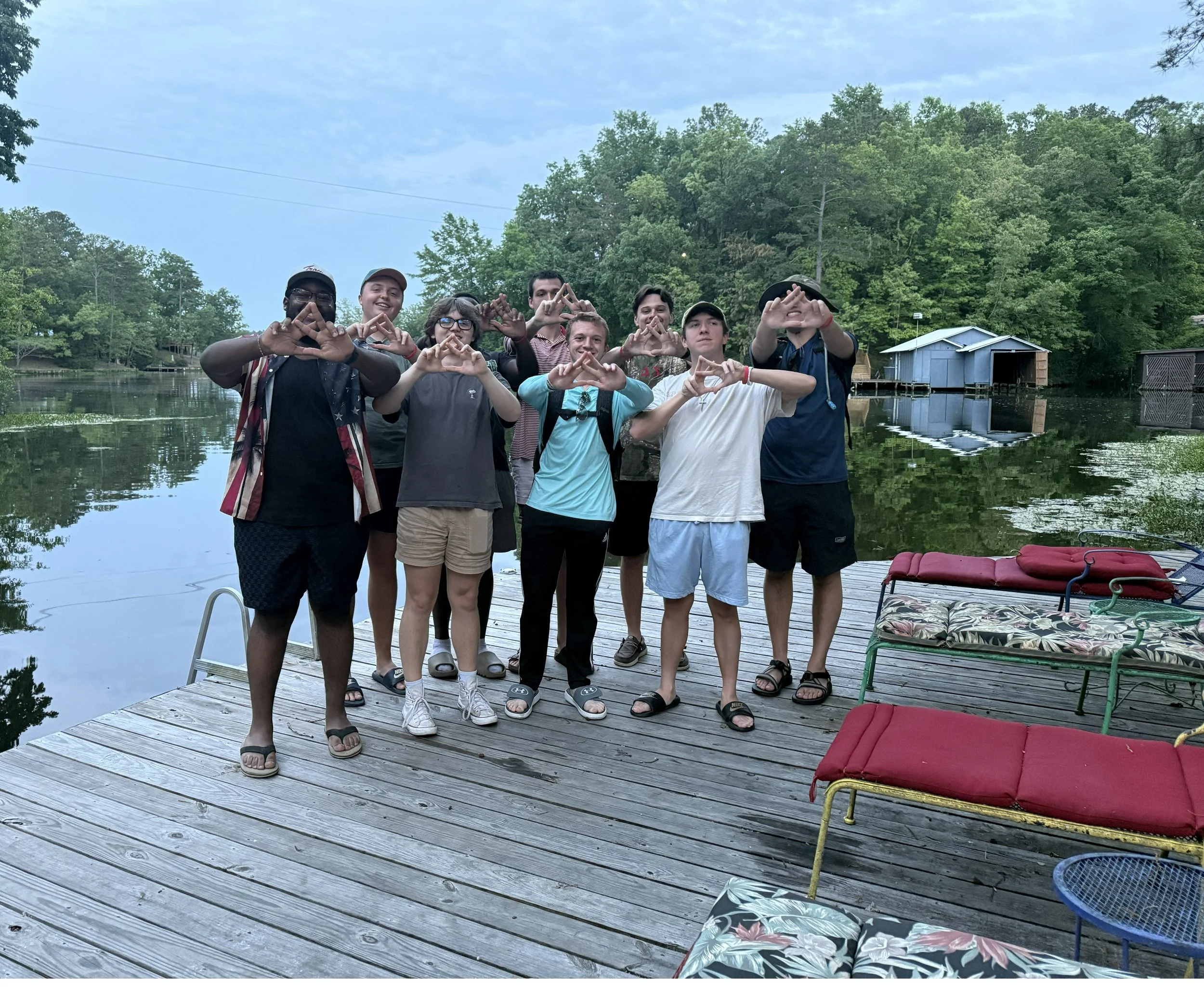 A group of nine young people standing on a wooden dock by a lake, making triangle hand gestures, with green trees in the background and some red and floral lounge chairs in the foreground.