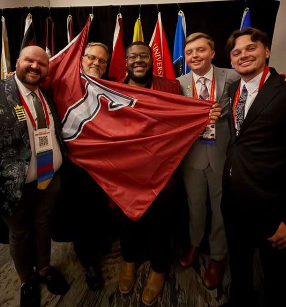 Group of five men at a conference, holding a red and white flag, with multiple international flags in the background.