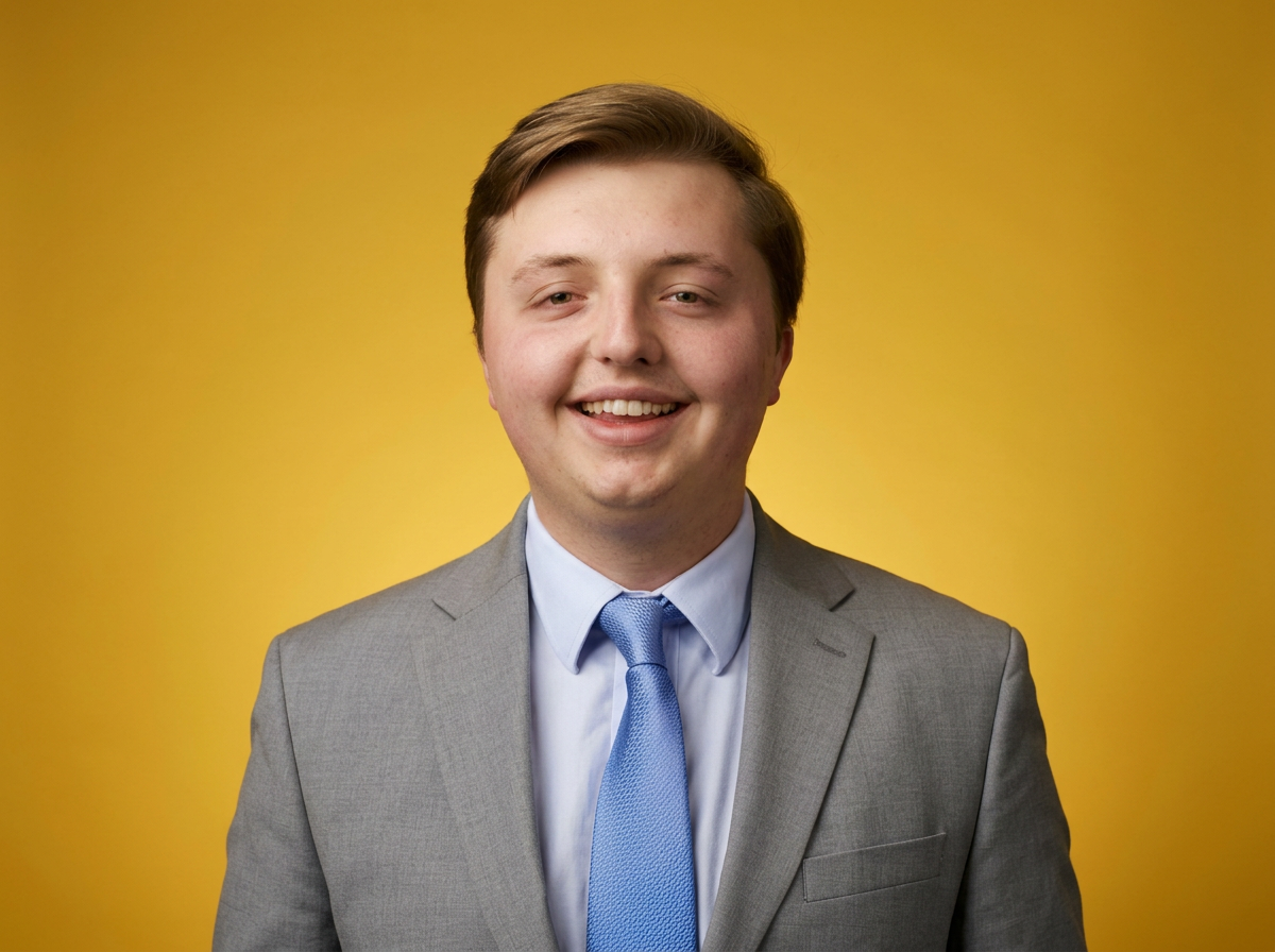 Young man in gray suit with blue tie smiling in front of a yellow background.