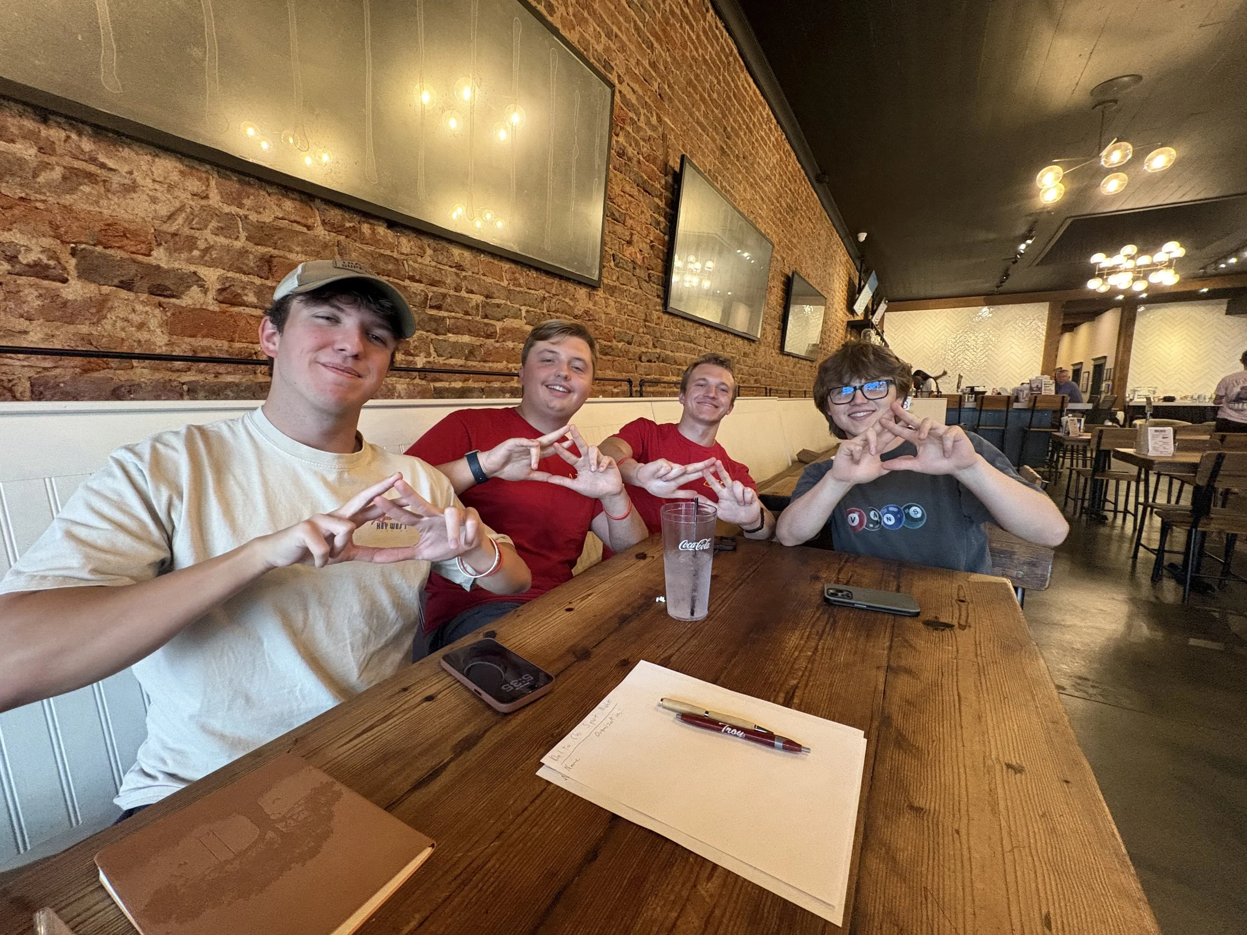 Four young men sitting at a wooden table in a restaurant, smiling and making triangular hand signs. The table has a notebook, a pen, a smartphone, and a glass of Coca-Cola. The background has a brick wall with framed mirrors and warm lighting fixtures.