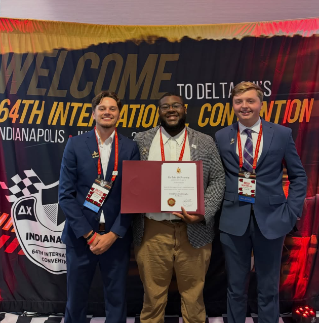Three young men in suits standing together at an awards ceremony, with a certificate in hand, in front of a backdrop that reads 64th International Convention, Indianapolis.
