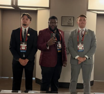 Three men in formal suits taking a mirror selfie at a conference, each wearing conference badges around their necks.