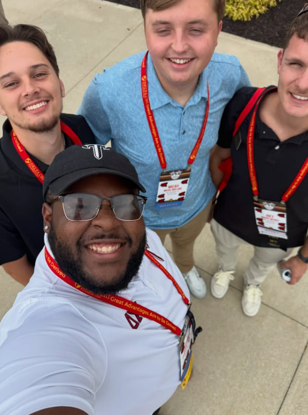 Four young men smiling and taking a selfie outdoors during an event, all wearing event badges and lanyards.
