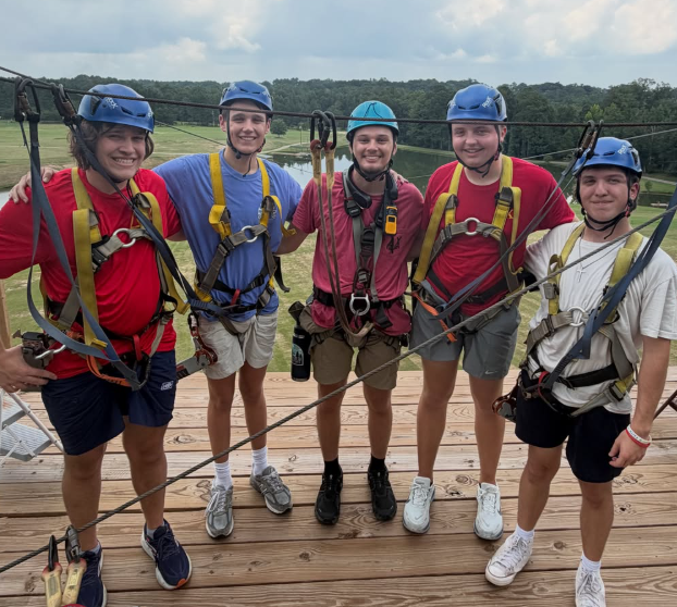 Five young people wearing safety harnesses and helmets stand on a wooden platform outdoors, prepared for zip-lining.