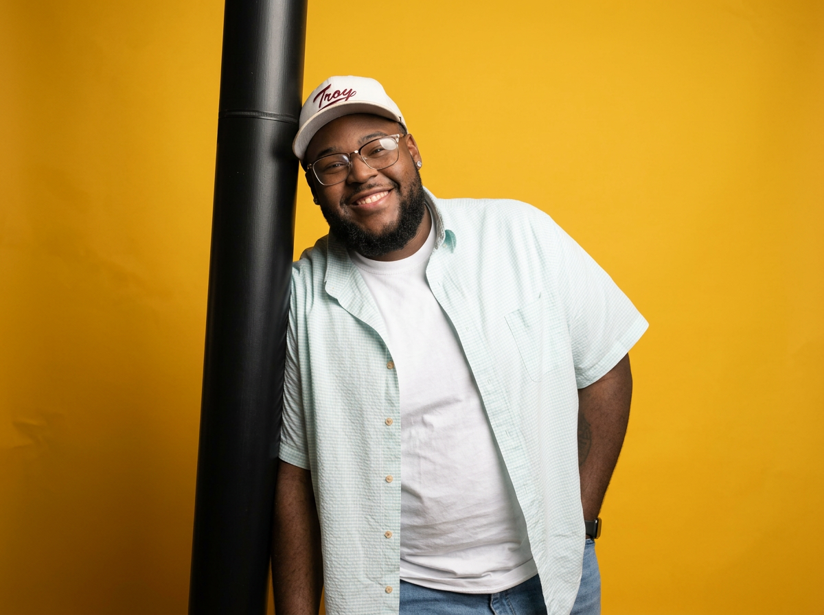 A smiling man with glasses, a beard, and earrings, leaning against a black pole with a yellow background. He is wearing a white cap with red writing, a light green button-up shirt over a white t-shirt, and has a tattoo on his left arm.