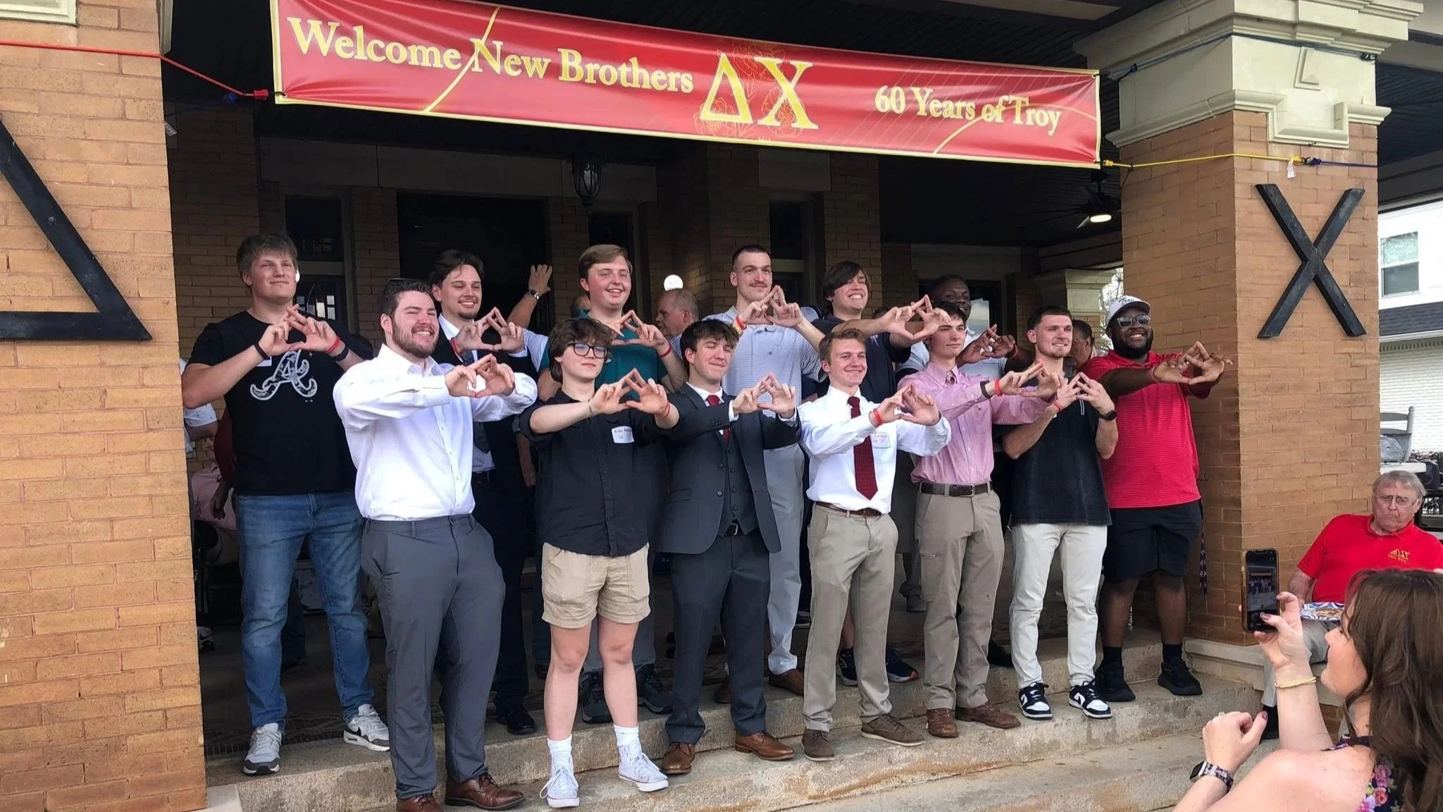 A group of young men, some in suits, standing on a porch, making triangle hand signs, celebrating a fraternity event under a banner that says 'Welcome New Brothers AX 60 Years of Troy'.