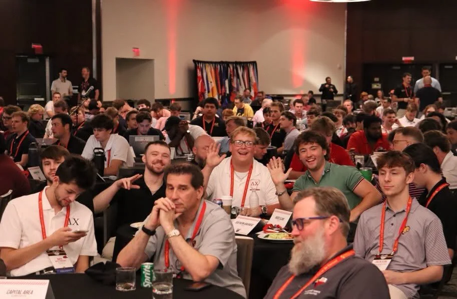 Large group of young men and boys seated at tables in a conference room, many wearing conference badges. Some are engaging with their phones, while others are smiling and waving at the camera. In the background, there are colorful flags and several people standing.