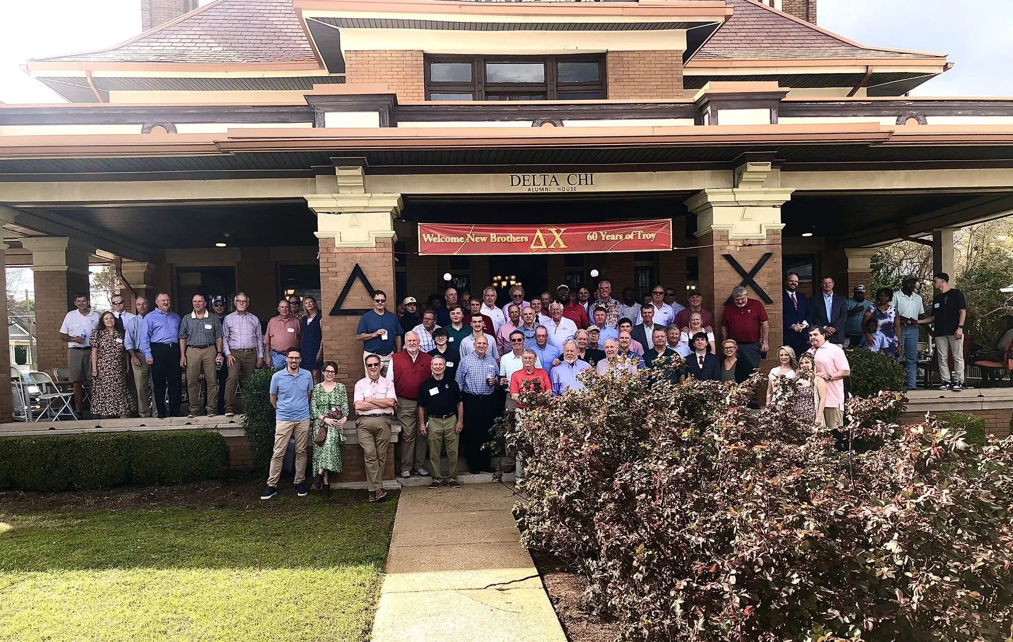 A large group of people gathered on the steps in front of a brick building with the sign 'Delta Chi Alumni House.' They are attending a reunion event, as indicated by the red banner that reads 'Welcome New Brothers, 60 Years of Troy.' The group includes men and women of various ages, dressed in casual and semi-formal clothing.