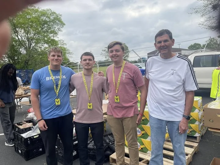 Four people standing outdoors, three young men and one older man, wearing medals around their necks, with a cloudy sky and parking lot in the background.