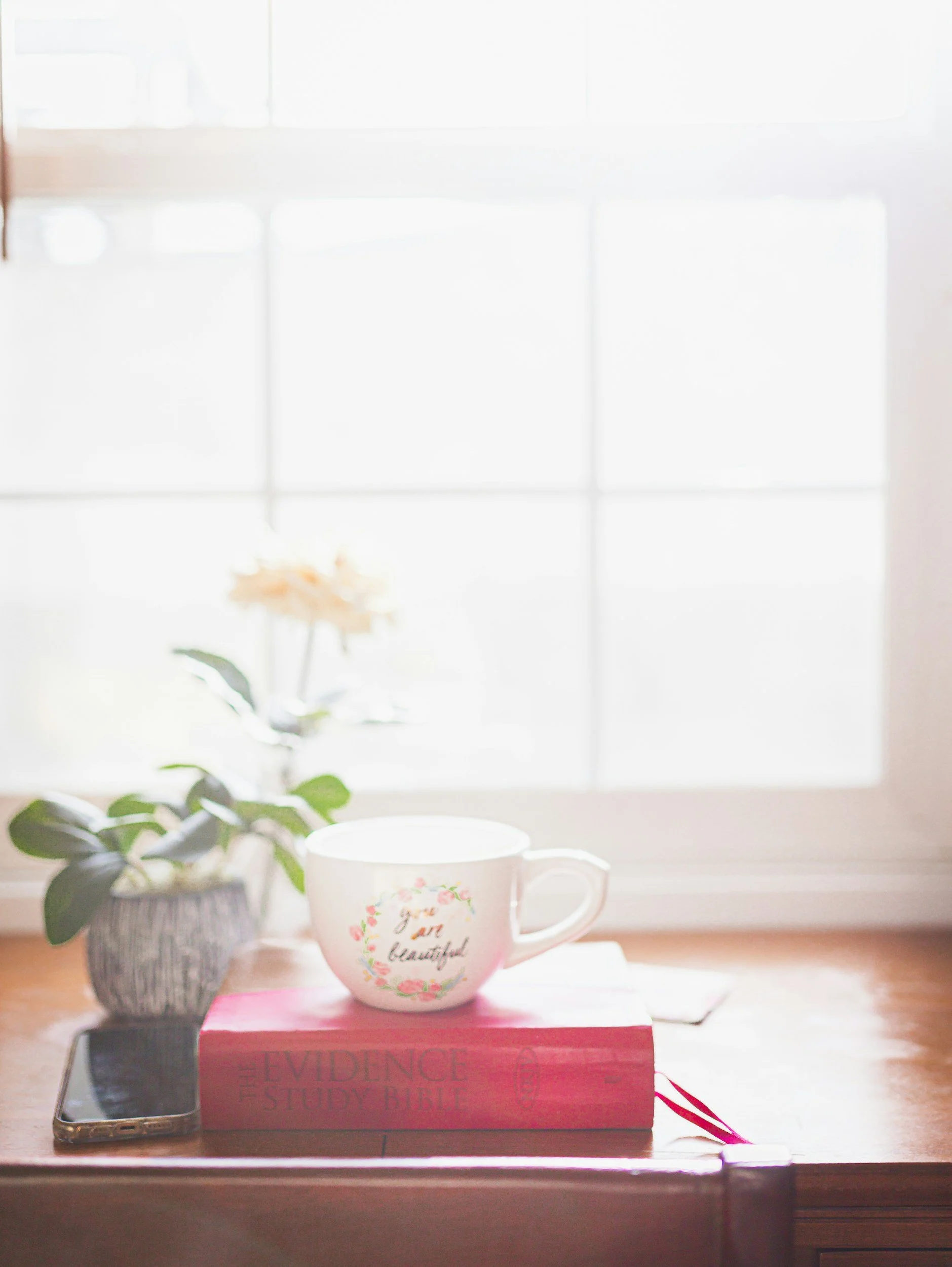 A white cup with floral decorations and the words 'You are so beautiful' printed on it, placed on a pink box labeled 'EVIDENCE STUDY BIBLE' on a wooden table, with a potted plant and a smartphone nearby, in front of a bright window.
