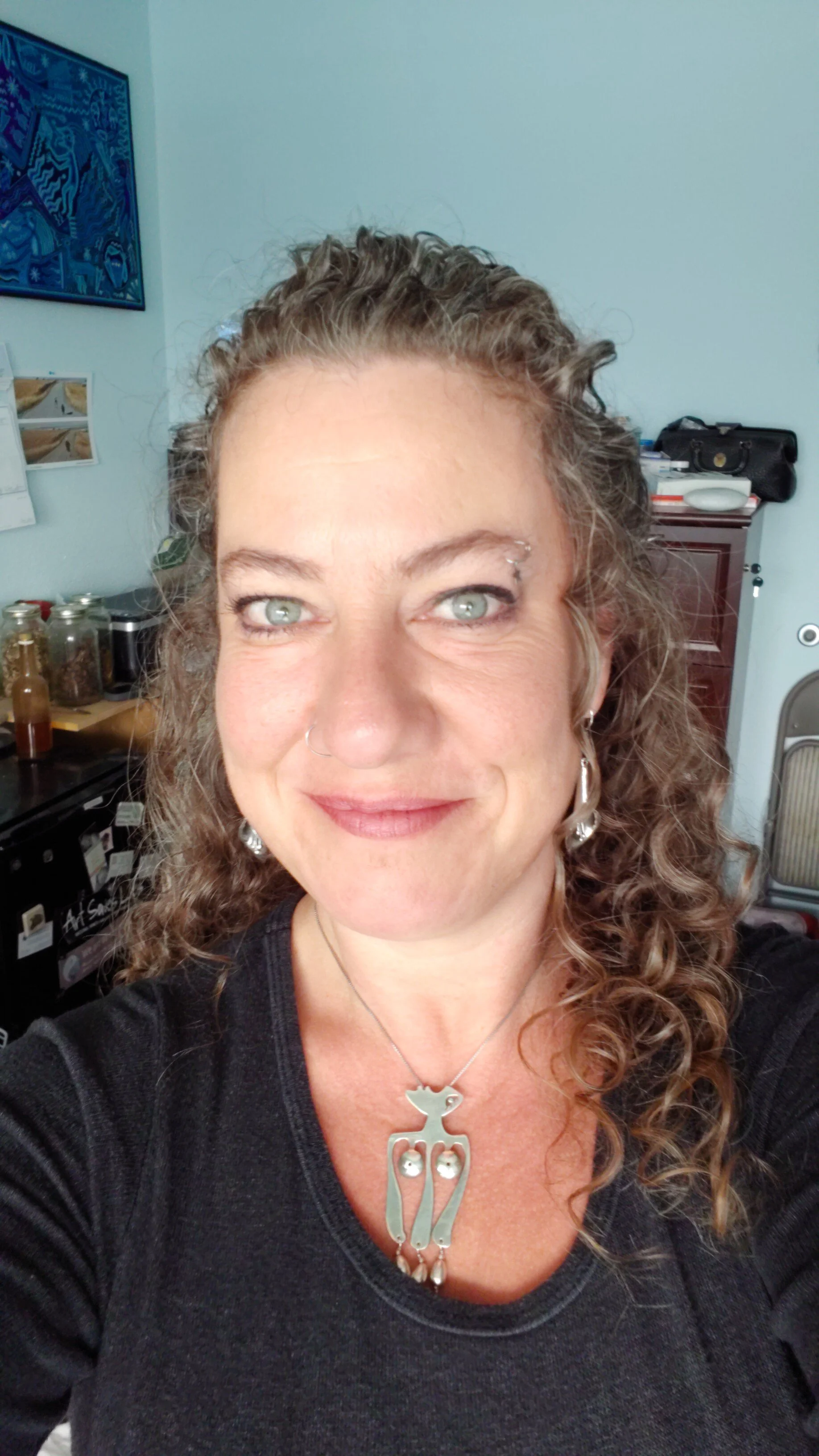 A woman with curly hair and light green eyes smiling, wearing earrings and a necklace with a cat-shaped pendant, taking a selfie in a room with various objects and artwork in the background.