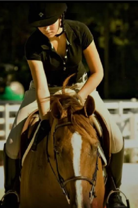 A woman riding a chestnut horse outdoors, wearing a black shirt, riding helmet, and beige riding pants.