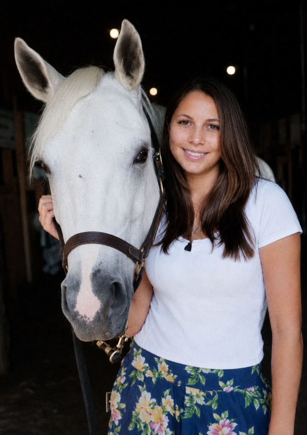 A woman with long dark hair smiling and posing with a white horse in a stable.