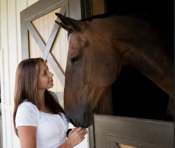 A woman with brown hair wearing a white shirt standing near a horse in a stable, touching the horse's nose and looking at it.