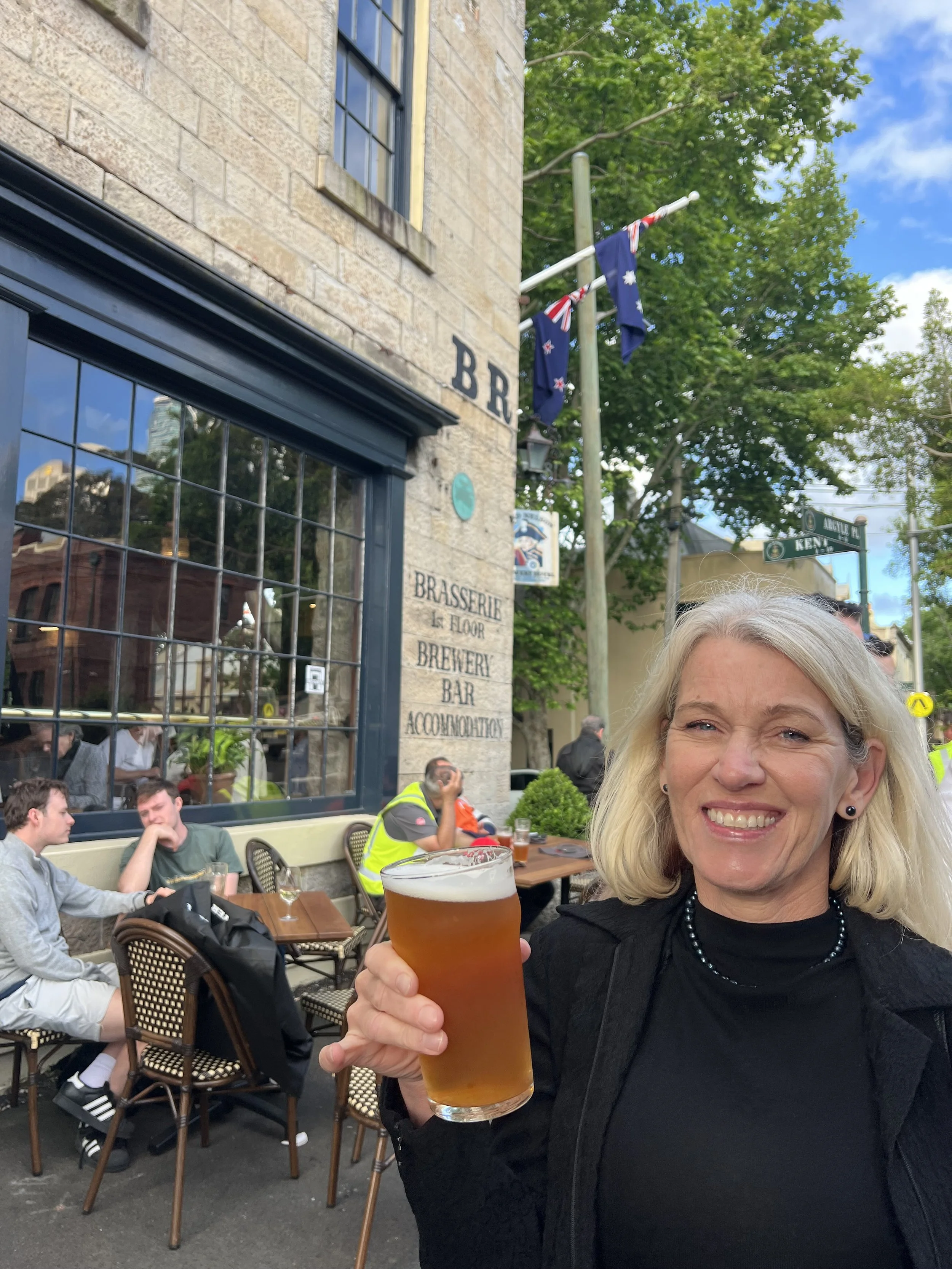 About us, our co-owner woman smiling and holding a pint of beer at an outdoor patio of a bar or restaurant.
