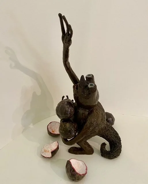 A dark, whimsical sculpture of a passionfruit grabbler creature with a long tail, holding and balancing three round passionfruits in front of a plain white background. The sculpture casts a shadow on the wall. Commission.
