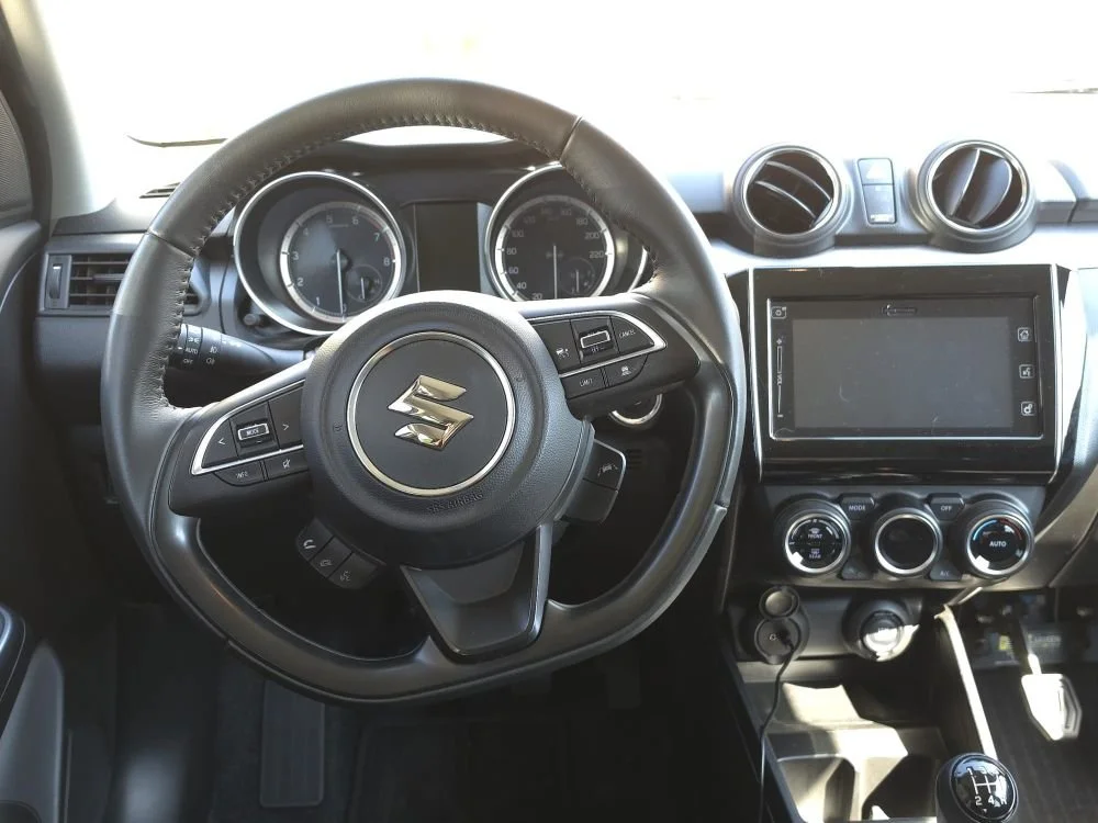 Interieur van een Suzuki-auto, inclusief stuurwiel, snelheidsmeter, touchscreen display, en klimaatregeling.