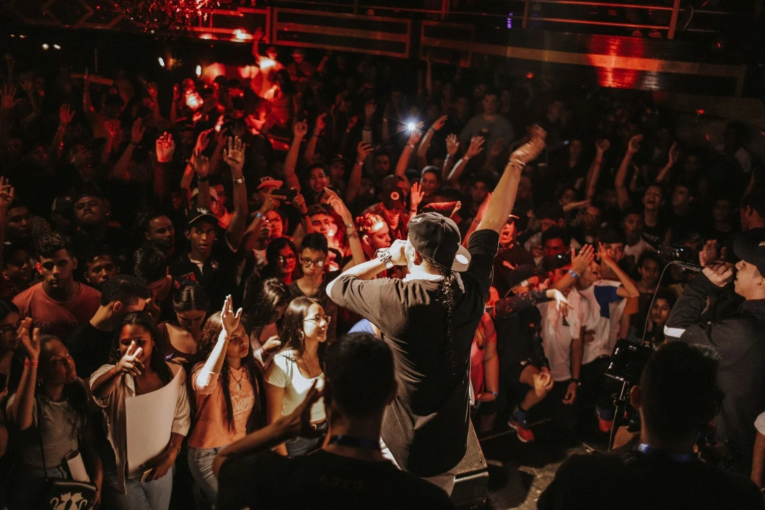 A concert with a performer on stage and an audience in front of them, some of whom are raising their hands and taking photos.