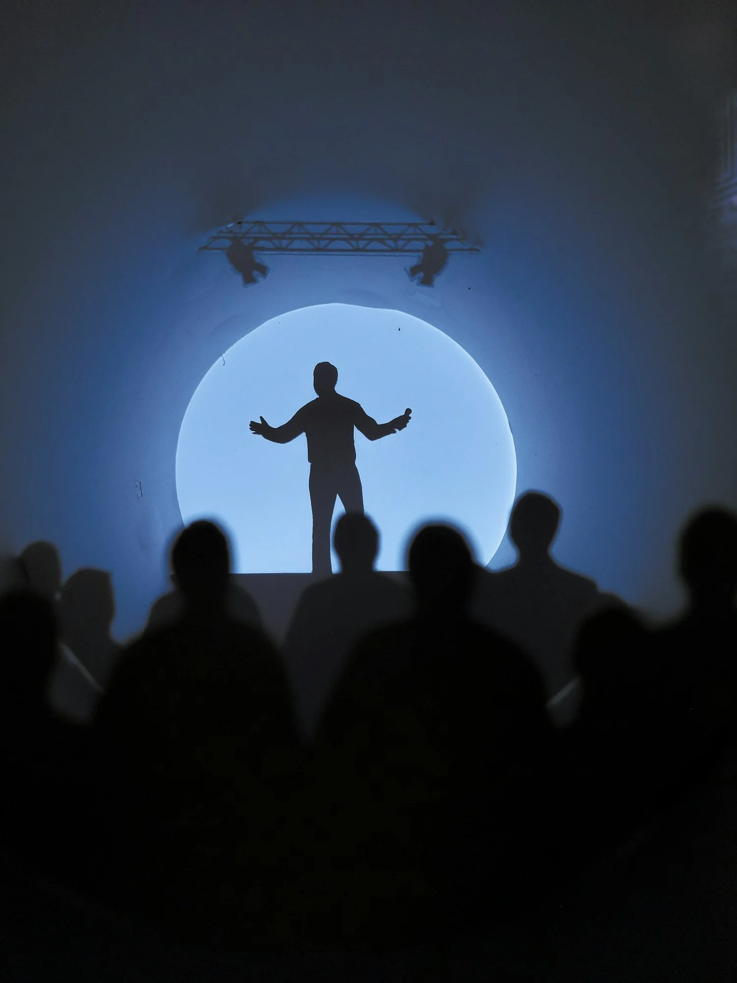 A person standing with arms outstretched in front of a large illuminated circular screen or tunnel, with audience members visible in the foreground and stage lights overhead.