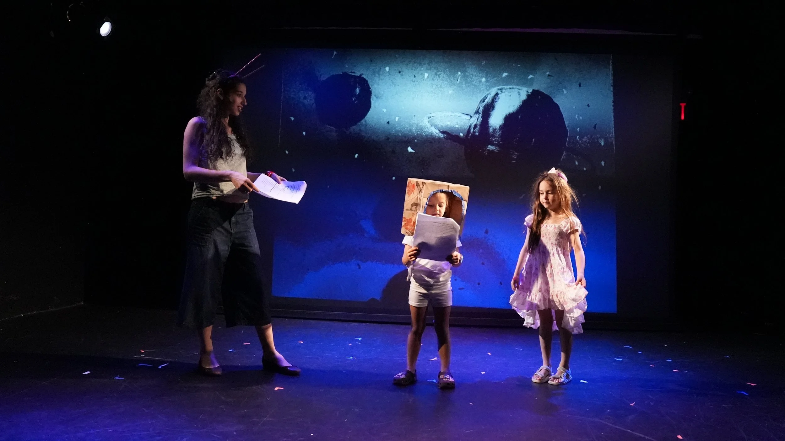 Three girls performing on a stage with a space-themed backdrop, one holding a script, one with a cardboard box on her head, and one in a pink dress.