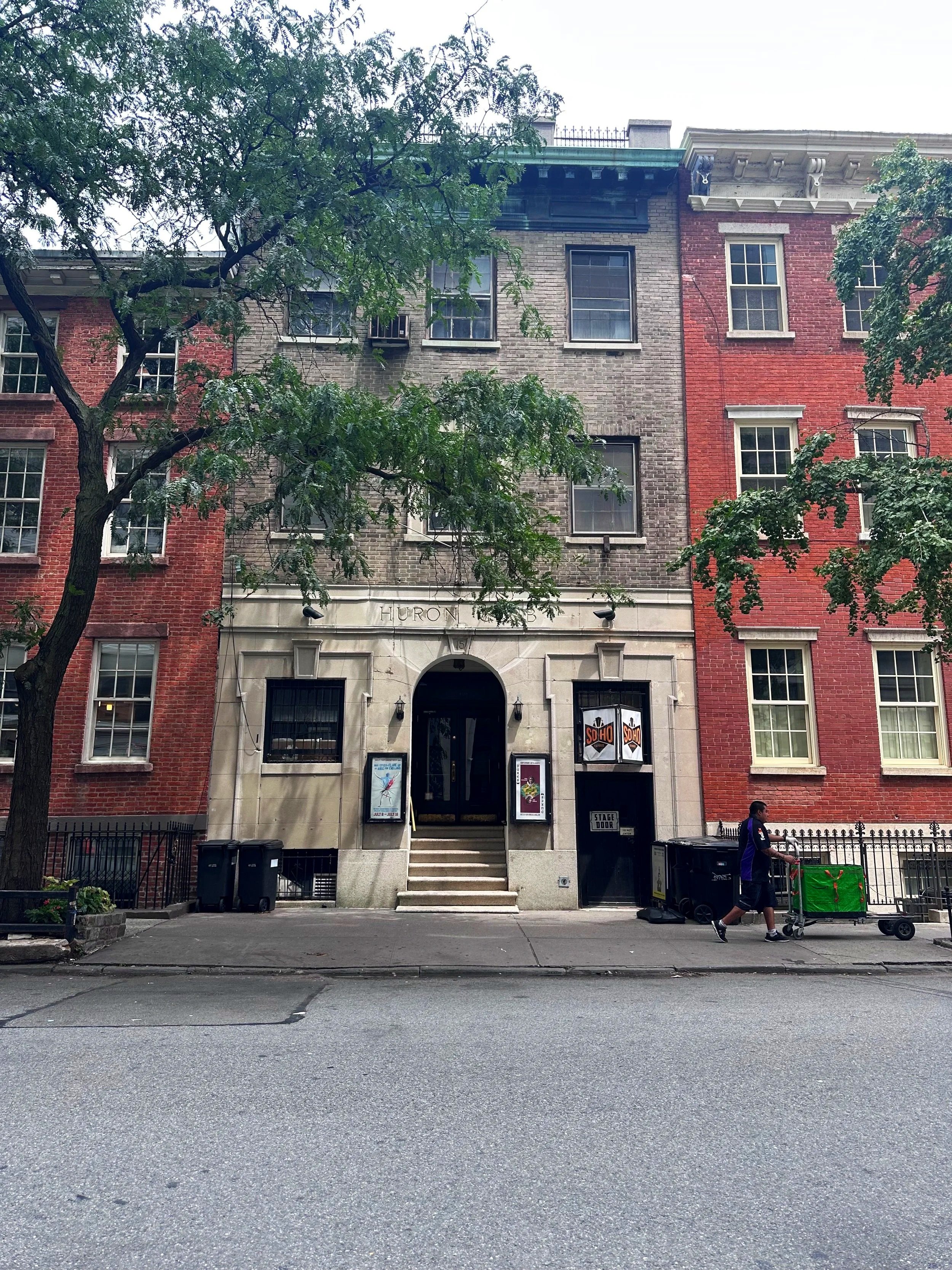 A city street scene featuring a brick building with an arched entrance, several multi-pane windows, and trees lining the sidewalk. A man is pushing a green cart or stroller, and black trash bins are in front of the building.