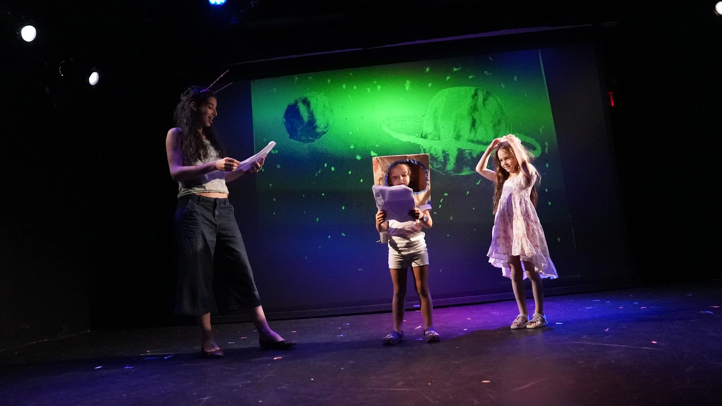 Three girls on a stage with a space-themed background, one holding papers, one wearing a cardboard space helmet, and one in a pink dress.